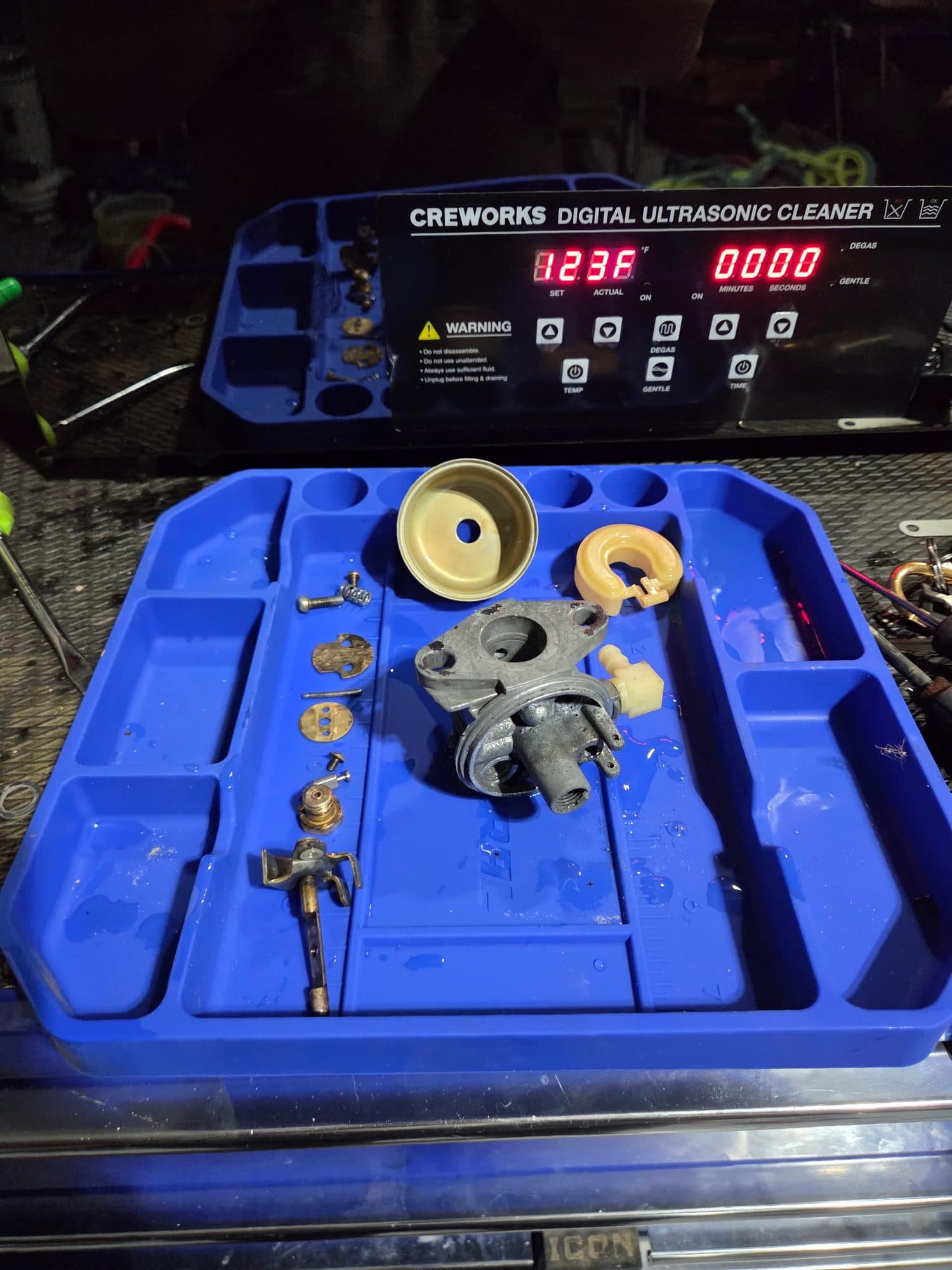 Carburetor parts laid out on the tray, the ultrasonic cleaner lit behind them