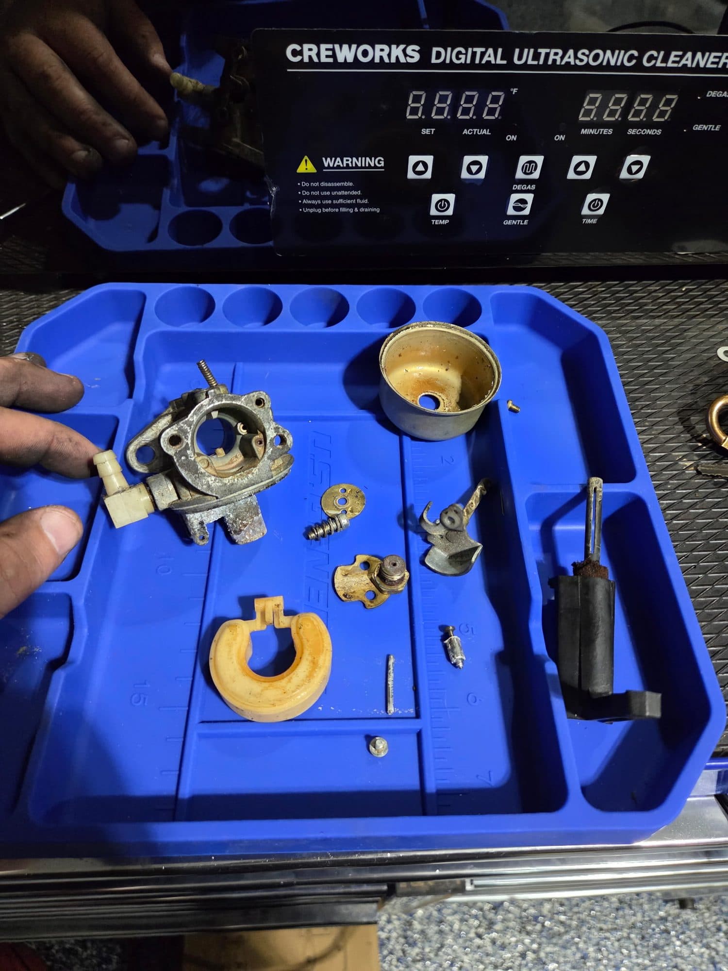A carburetor taken apart on a blue parts tray in front of the ultrasonic cleaner